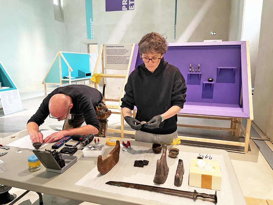 Photo du montage de l'exposition temporaire Bronze, l'âge retrouvé au Musée Savoisien à Chambéry : deux personnes travaillant au soclage des objets